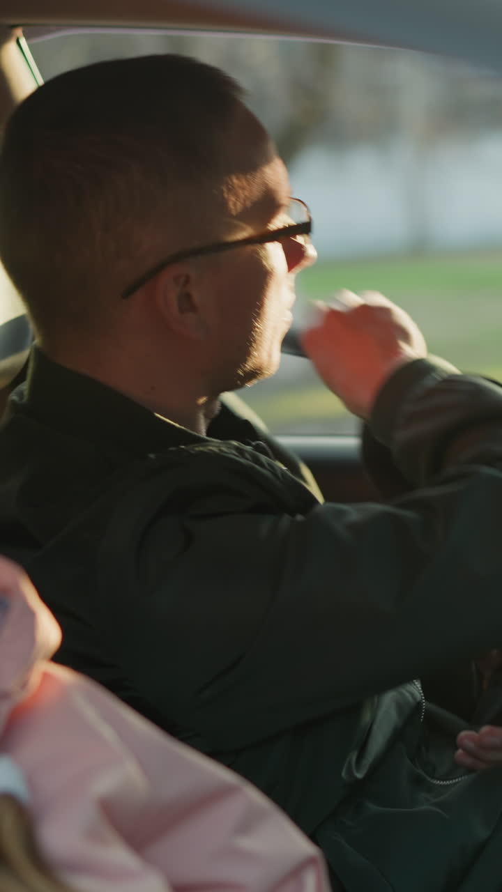 un hombre con una chaqueta verde se sienta en el asiento del conductor, sosteniendo un teléfono y acabando de desabrocharse el cinturón de seguridad. una joven con un sombrero de punto rosa y una bufanda blanca se sienta pacíficamente, mirando por la ventana