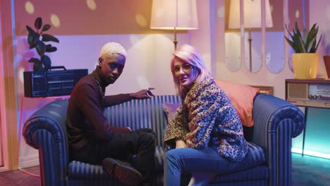 african american man and caucasian stylish woman in sunglasses talking while sitting on couch in a retro party at home