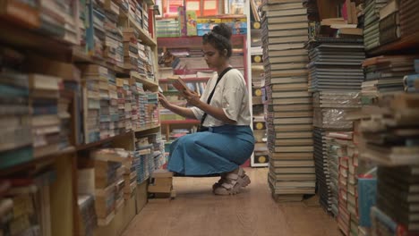 an attractive asian female reading the blurb on the back of a hardcover book in the isle of a bookstore, india