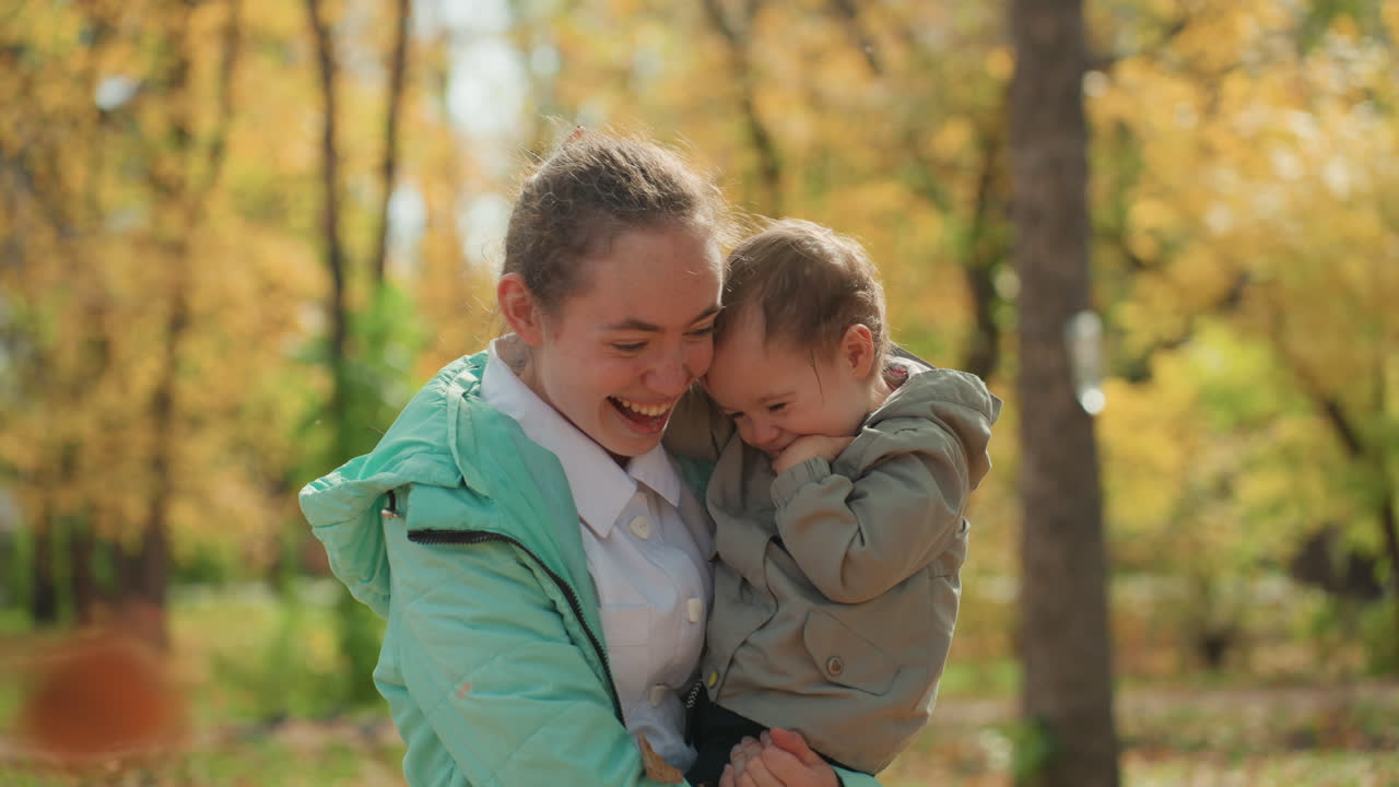 Mutter und Kleinkind spielen inmitten fallender Blätter im Park, fröhliches Lachen, strahlendes Lächeln, spielerisches Werfen von Laub, grüne Jacke, gemütlicher Mantel, goldener Baumbestand, authentisches Familienglück, lebhafte Herbstenergie.