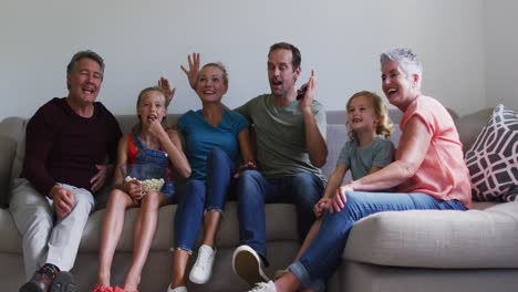 excited caucasian three generation family sitting on couch watching tv, eating popcorn and cheering