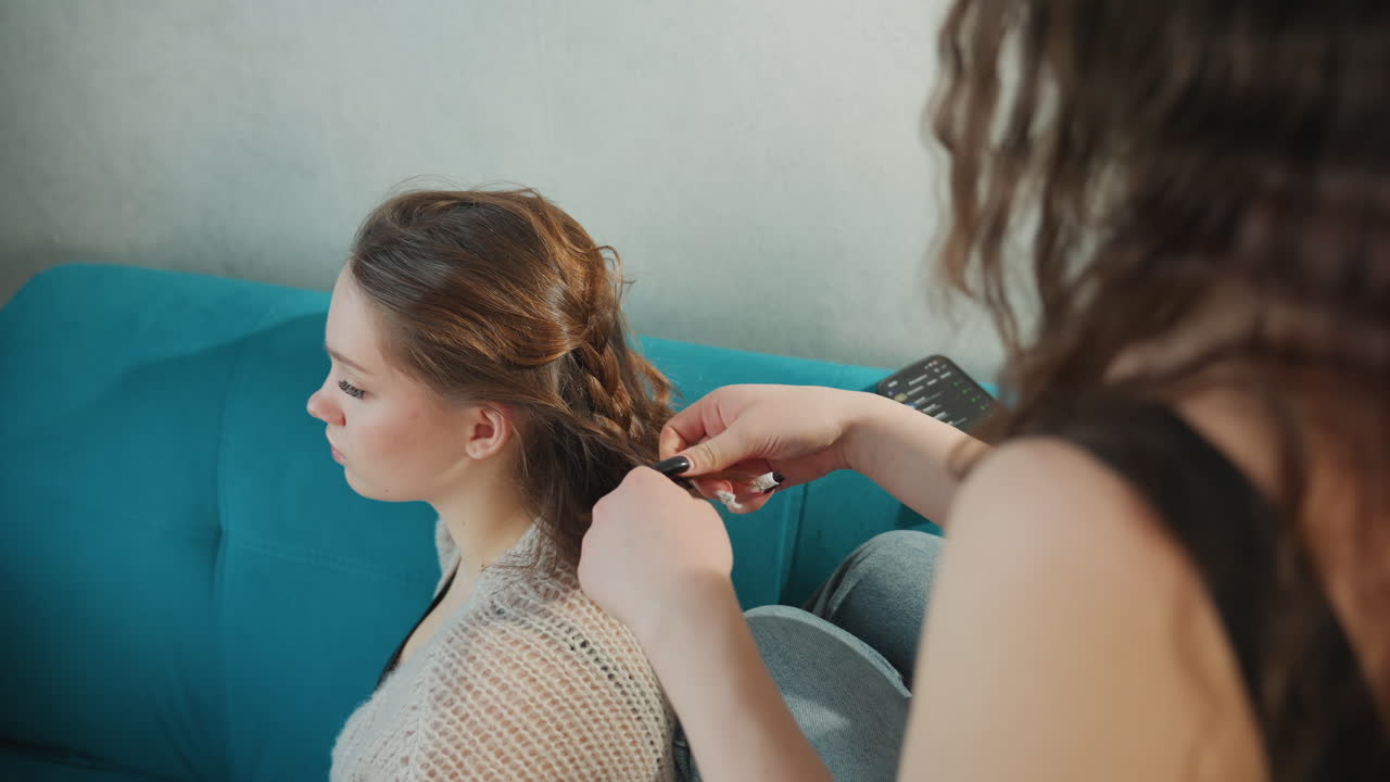 Jovem com tranças no cabelo, mulher sentada em um sofá azul-petróleo enquanto a amiga faz uma trança elaborada; garota sentada no sofá enquanto sua amiga cria cuidadosamente um penteado complexo com atenção concentrada
