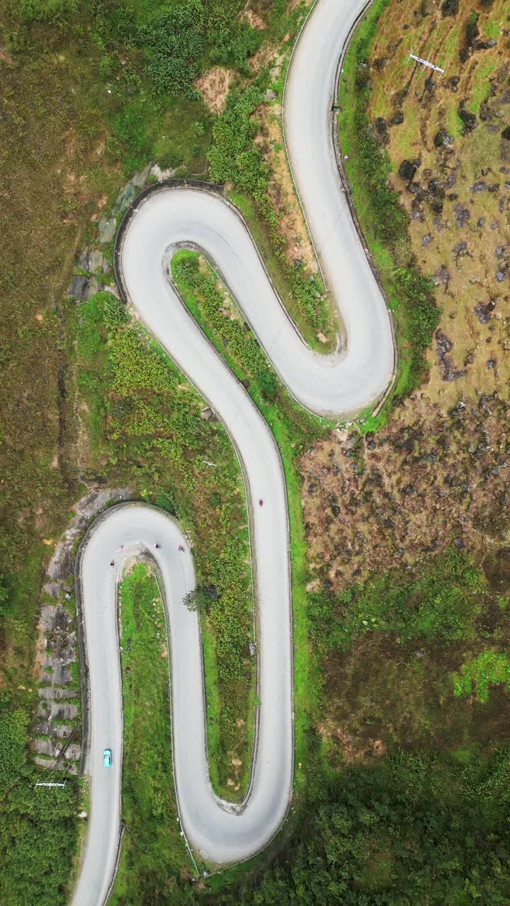 Vertikalt dronebilde ovenfra av en fjellvei med skarpe hårnålssvinger langs en grønn dalside, med motorsykler som kjører gjennom ulendt terreng.