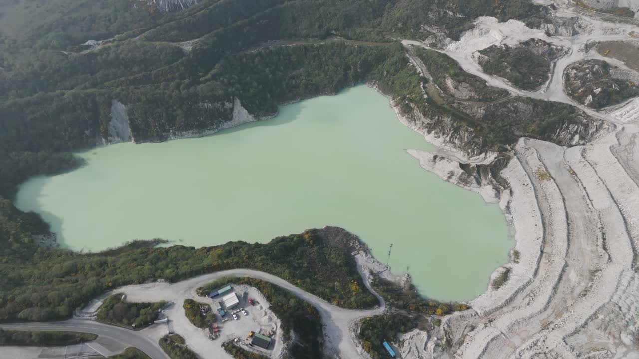 Oblique aerial of milky turquoise lake filling abandoned quarry, encircled by stepped excavation scars and sparse vegetation, illustrating post-industrial land regeneration