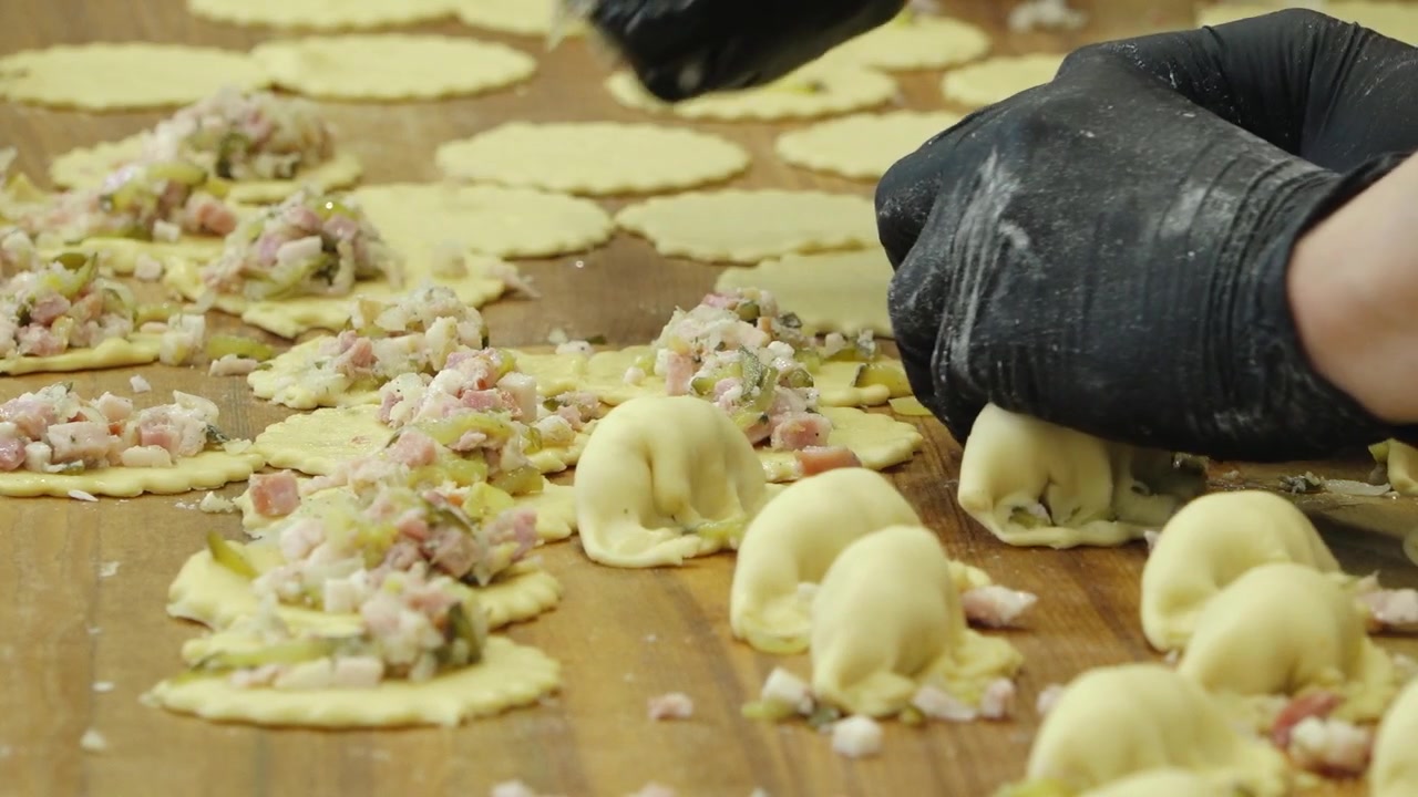 Mano guantata di nero piega l'impasto ripieno per pasticceria su una superficie di lavoro in legno con ripieno salato e pezzi di impasto preparati in una cucina di panetteria.