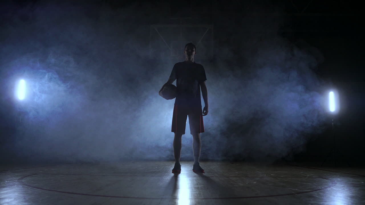 Karanlık bir arka planda, basketbol sahasında dumanlar içinde bir basketbol oyuncusunun silueti, ağır çekimde topu atıyor ve kameraya bakıyor.