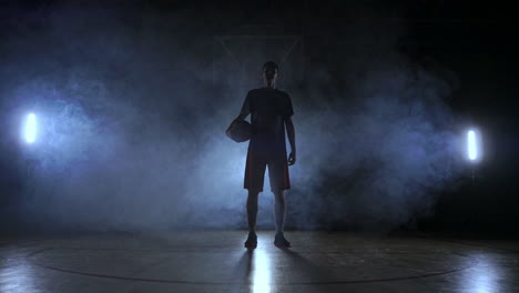 Die-Silhouette-Eines-Basketballspielers-Auf-Dunklem-Hintergrund-Mit-Rauch-Auf-Dem-Basketballplatz-Wirft-Einen-Basketball-Und-Blickt-In-Zeitlupe-In-Die-Kamera