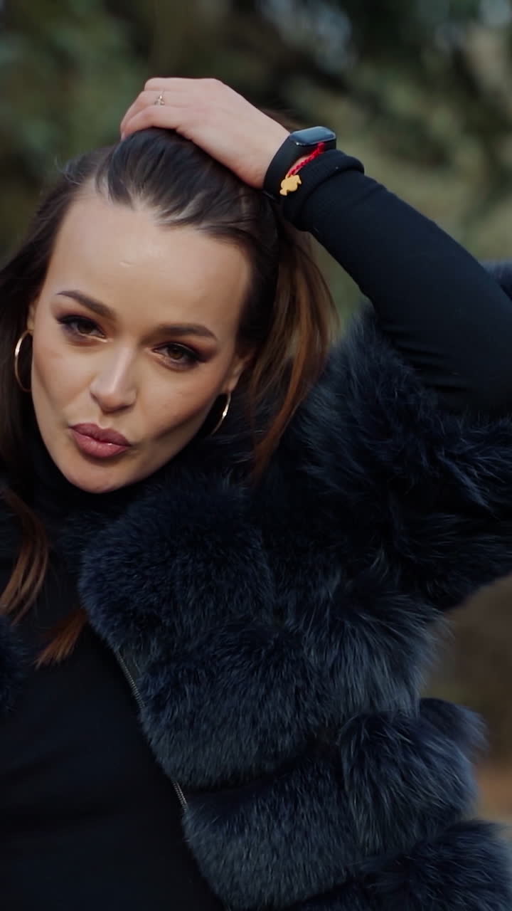 Portrait of a fashionable girl outdoors. Pretty-looking brunette model posing on camera in fur coat with a cup of coffee in the park. Slow motion. Vertical video