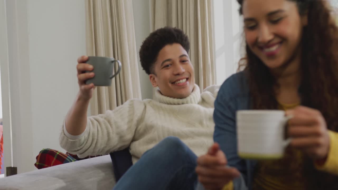 Video of happy biracial couple siting on sofa under blanket and drinking coffee