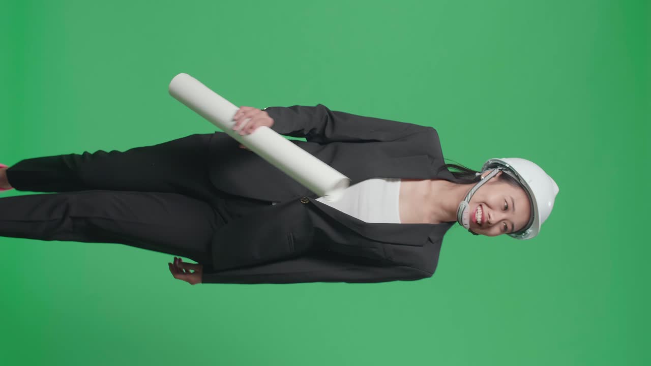 Smiling Asian Female Engineer With Blueprint Looking Around And Shaking Her Head While Walking In The Green Screen Studio