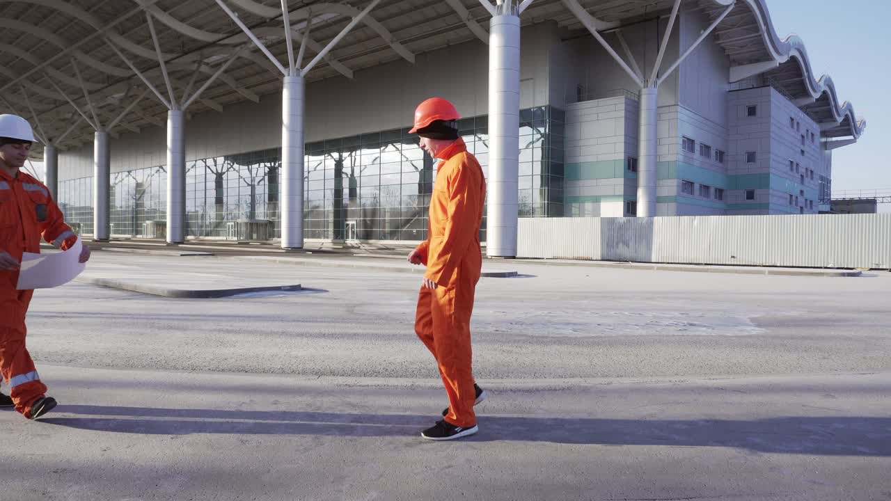 двое строителей в оранжевой форме и касках встречаются на строительном объекте, пожимают друг другу руки и вместе осматривают построенное здание