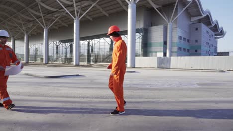 Two-Construction-Workers-In-Orange-Uniform-And-Helmets-Meeting-Each-Other-At-The-Bulding-Object,-Shaking-Hands-And-Examining-The-Constructed-Building-Together