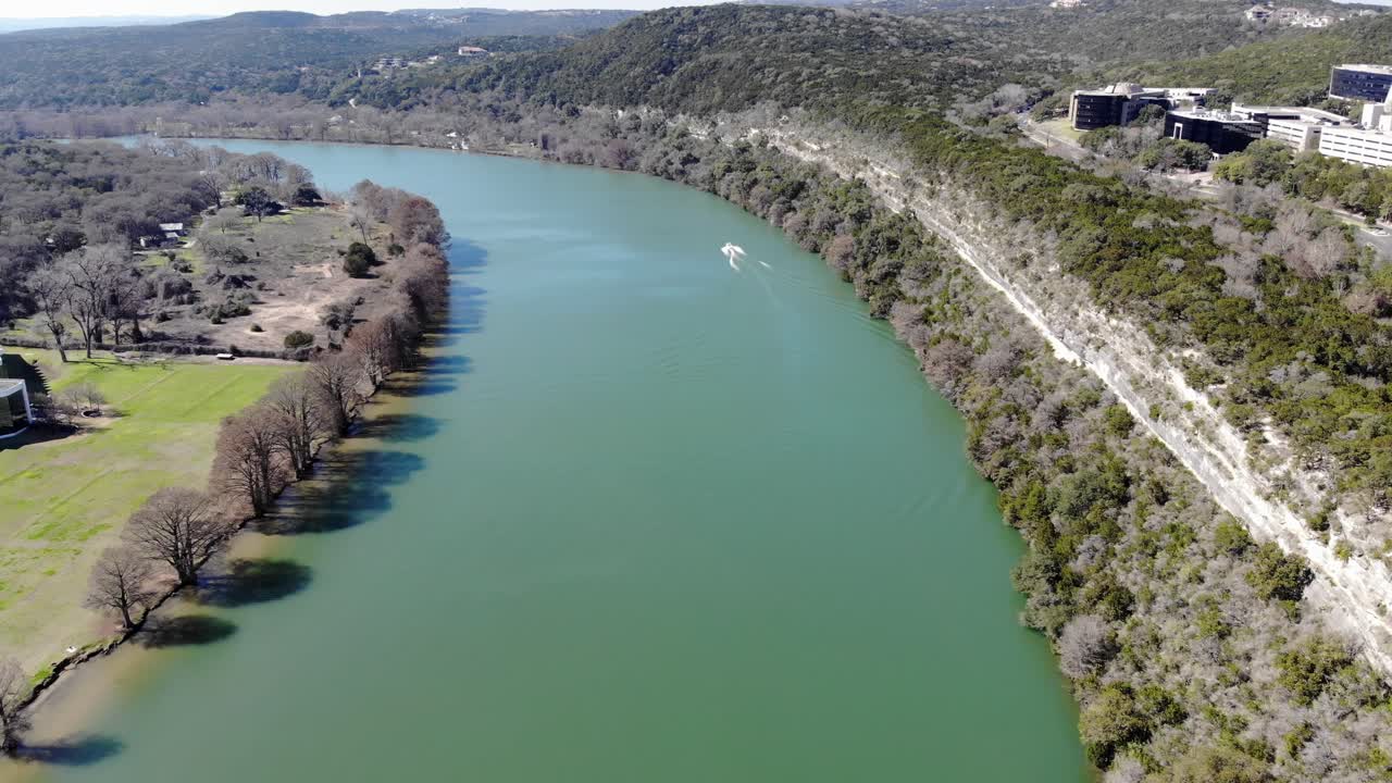 Aerial Austin Pennybacker Bridge - moving up river in the direction of boat. Great view of brightly colored water - sheer rock cliffs on the right. Ripples in the water visible from the boat's wake.