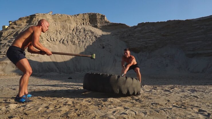 Two powerful male athletes training together hit the wheel with a hammer at sunset in the desert. Endurance training