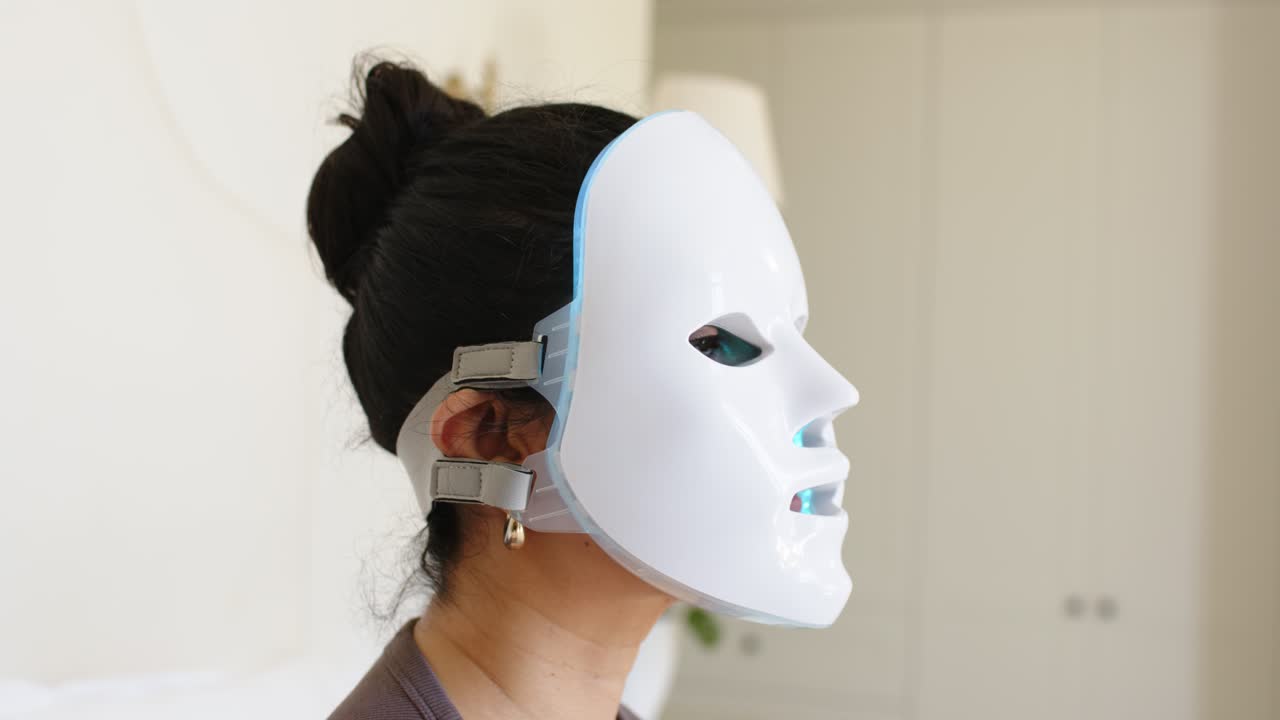 En kvinde løfter en LED-maske mod ansigtet derhjemme, justerer stropper, mens et blåt lys gløder til hudpleje.