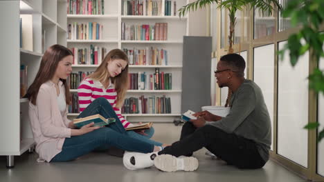 three students sitting on the floor in a library discussing books