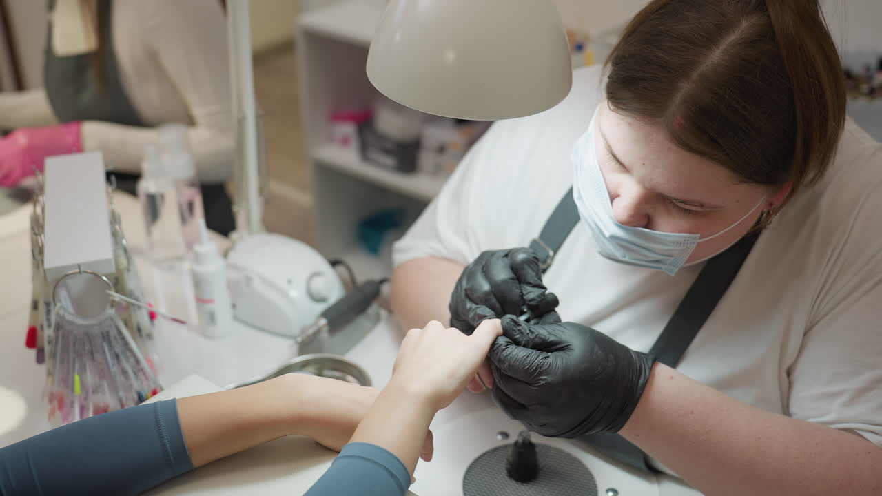 Nail technician in black glove and face mask carefully applying hardener on customer fingernail during manicure session under focused salon lighting with tools and nail samples visible on workspace