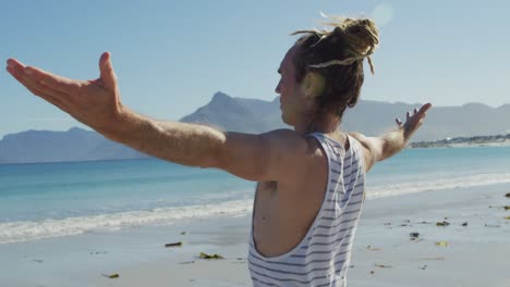 Video-Eines-Kaukasischen-Mannes-Mit-Dreadlocks,-Der-Am-Sonnigen-Strand-Mit-Ausgestreckten-Armen-Yoga-Praktiziert