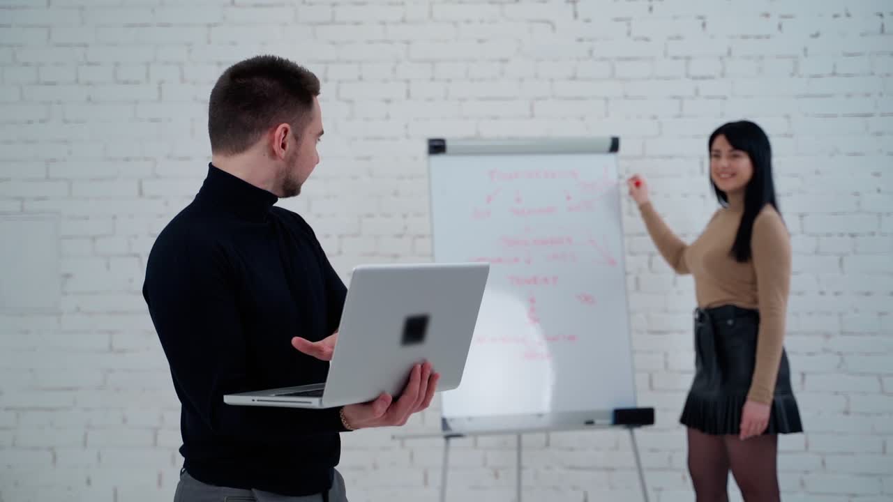 Young businessman and beautiful woman. Female partner writes business project on a board. Handsome man working on a laptop and approves woman's job.