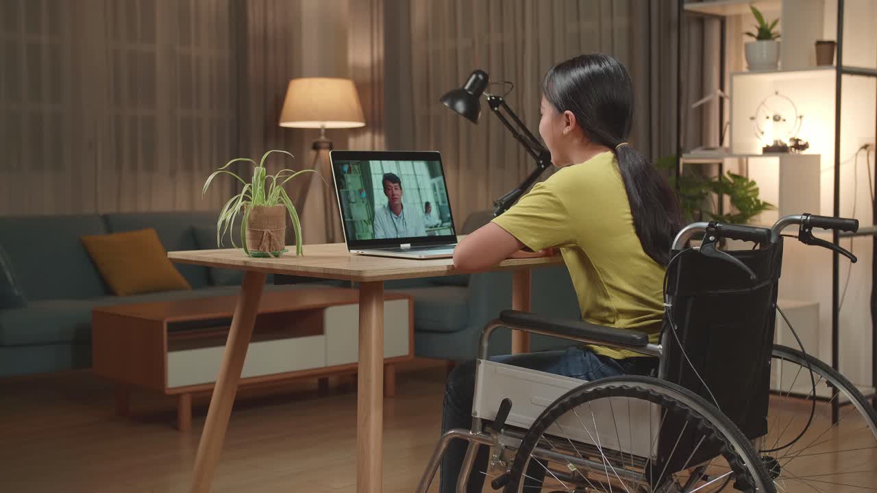 Back View Of Asian Kid Girl Sitting In A Wheelchair While Video Call With Doctor On Laptop Computer At Home