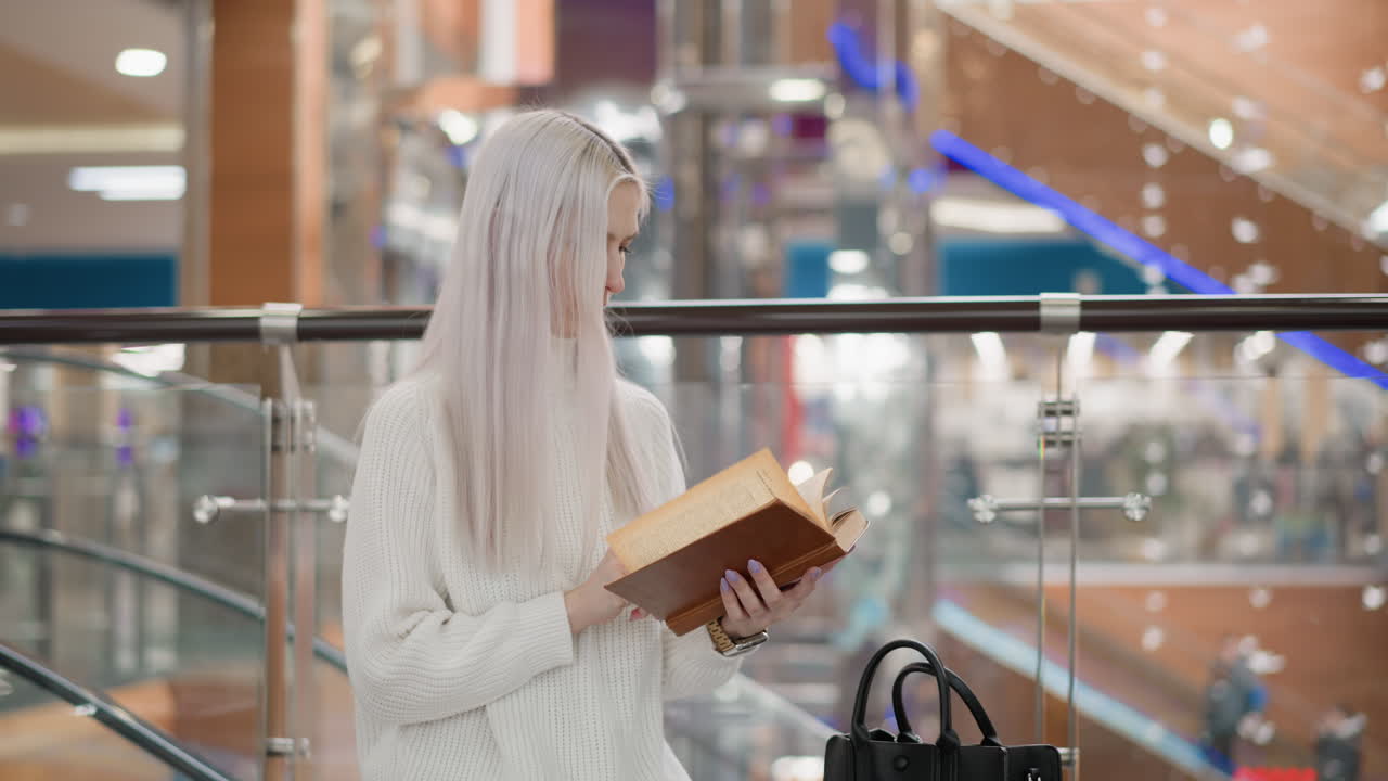 determined woman seated on mall bench flipping through pages of book with focused gaze wearing white sweater and jeans black bag beside modern glass railing escalator blurred background