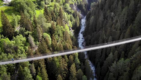 Aerial-view-alongside-of-the-Goms-suspension-bridge-with-a-hiker-walking-across-high-up-above-Rhone-river-valley-in-Valais,-Switzerland-high-up-above-the-river-and-trees