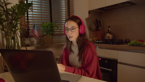 A-confident-brunette-girl-in-red-wireless-headphones-with-glasses-and-a-red-sweater-sits-at-a-table-in-the-kitchen-in-front-of-a-gray-laptop-and-studies-foreign-languages-near-her-the-flag-of-the-United-States-of-America-in-a-modern-apartment-in-the-kitchen