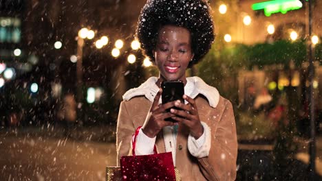 close-up view of african american woman holding shopping bag and using smartphone on the street while it‚äôs snowing in christmas