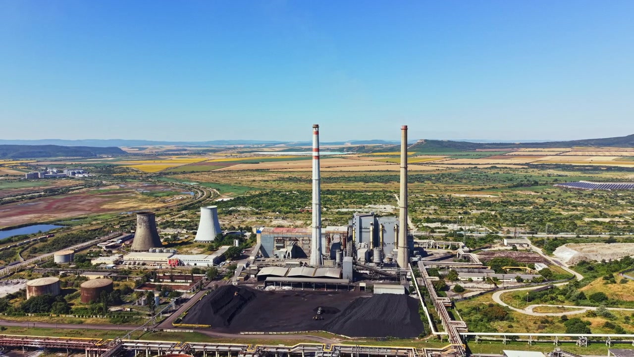 Centrale elettrica situata in un'area rurale con grandi torri di raffreddamento e ciminiere mostra attività industriale nel paesaggio sotto un cielo azzurro e limpido durante il giorno.