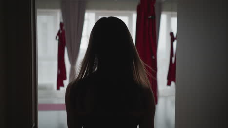 back view lady walking into room through doorway silhouette framed by door edges with natural light streaming from window red curtains in background capturing serene interior transition