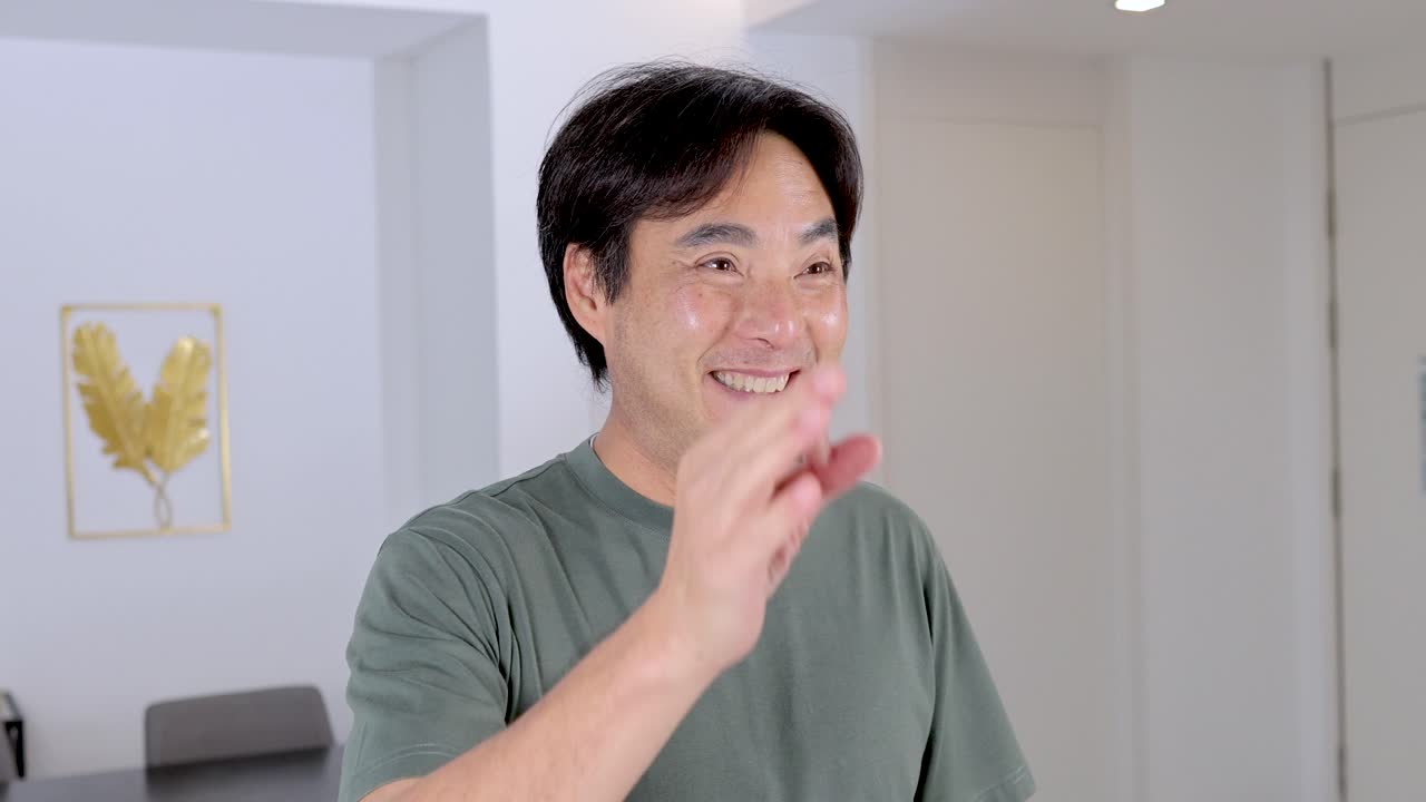 Asian man smiles confidently, making OK hand gesture in bright, modern indoor setting, steady camera