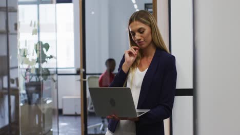 Smiling-caucasian-woman-using-laptop-while-standing-at-modern-office