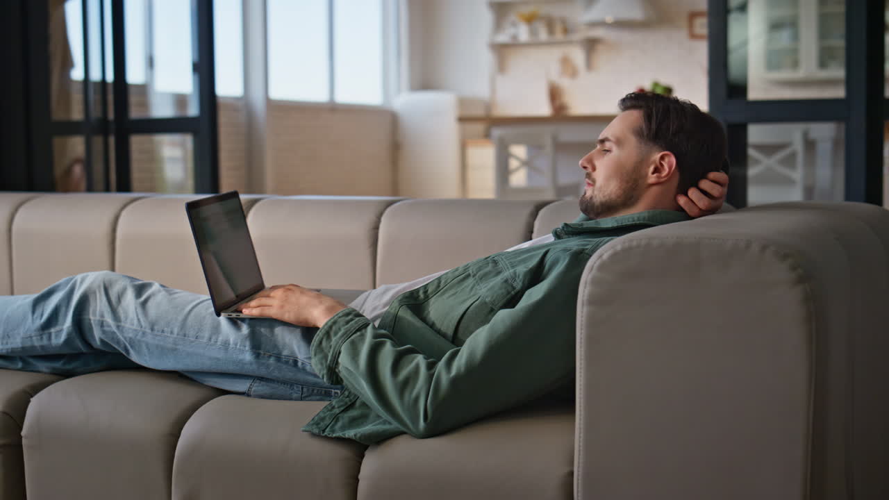 Chilling freelancer looking computer lying beige sofa. Relaxed man working couch