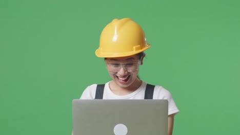 primer plano de una trabajadora asiática con gafas de protección y casco de seguridad mirando la computadora portátil y luego gritando gol celebrando mientras está de pie en la pantalla verde de fondo del estudio