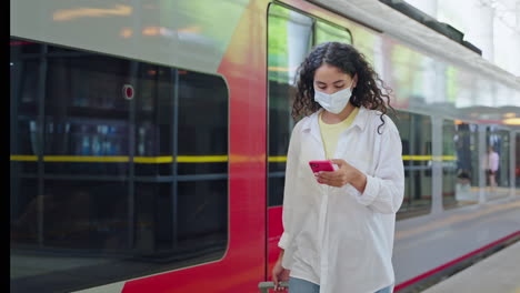 woman waiting at a train station, wearing a mask and using a phone