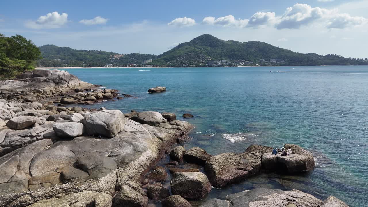 드론 영상은 태국 푸켓의 조용한 바위 해안선을 포착합니다. 은 파란 물과 먼 언덕은 빛 하늘 아래 있습니다.