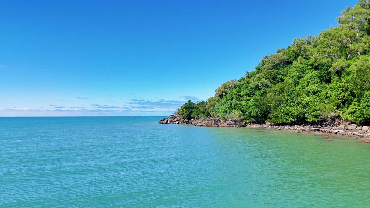 Drone pans over lush rainforest headland, turquoise ocean, rocky shore, under bright daylight sky