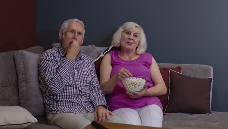 portrait of elderly couple watching tv at home eating popcorn enjoying film together at home