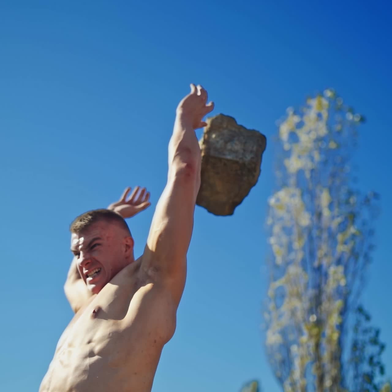 Bodybuilder is training outdoors. Strong athlete with muscular body rising heavy stone and throwing it over his head on the rocky background. Slow motion.