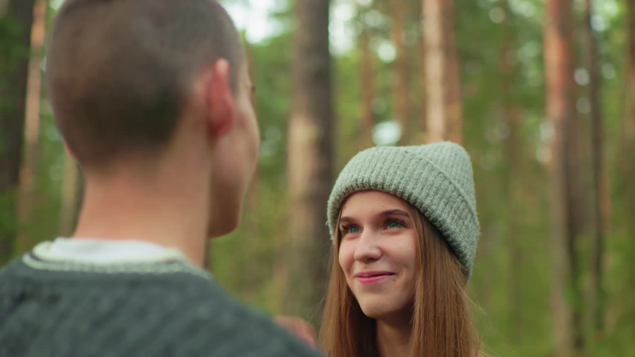 Girl approaches boy in forest, gently removing dirt from his eye as he playfully covers her face with head warmer, sharing a lighthearted and affectionate moment surrounded by trees