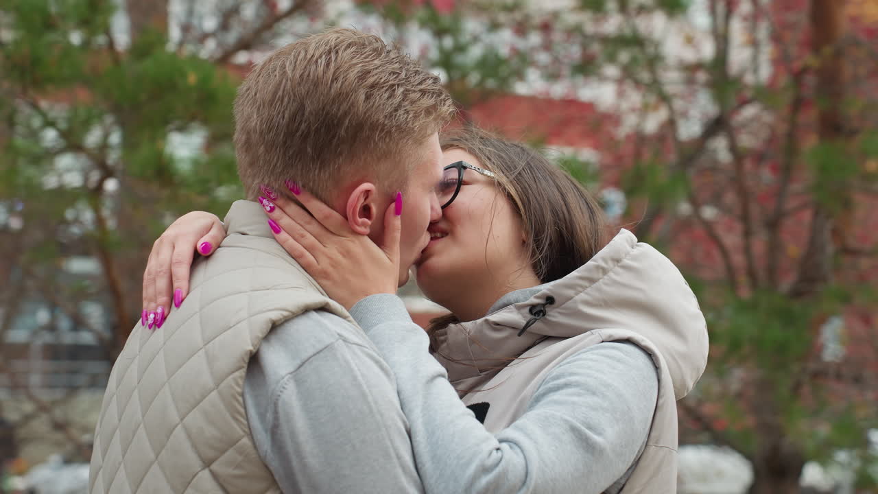 Lovers engross in intimate kiss outdoors wearing same outfit as wind gently sways hair and leaves around them, creating romantic moment with cars passing by in urban background