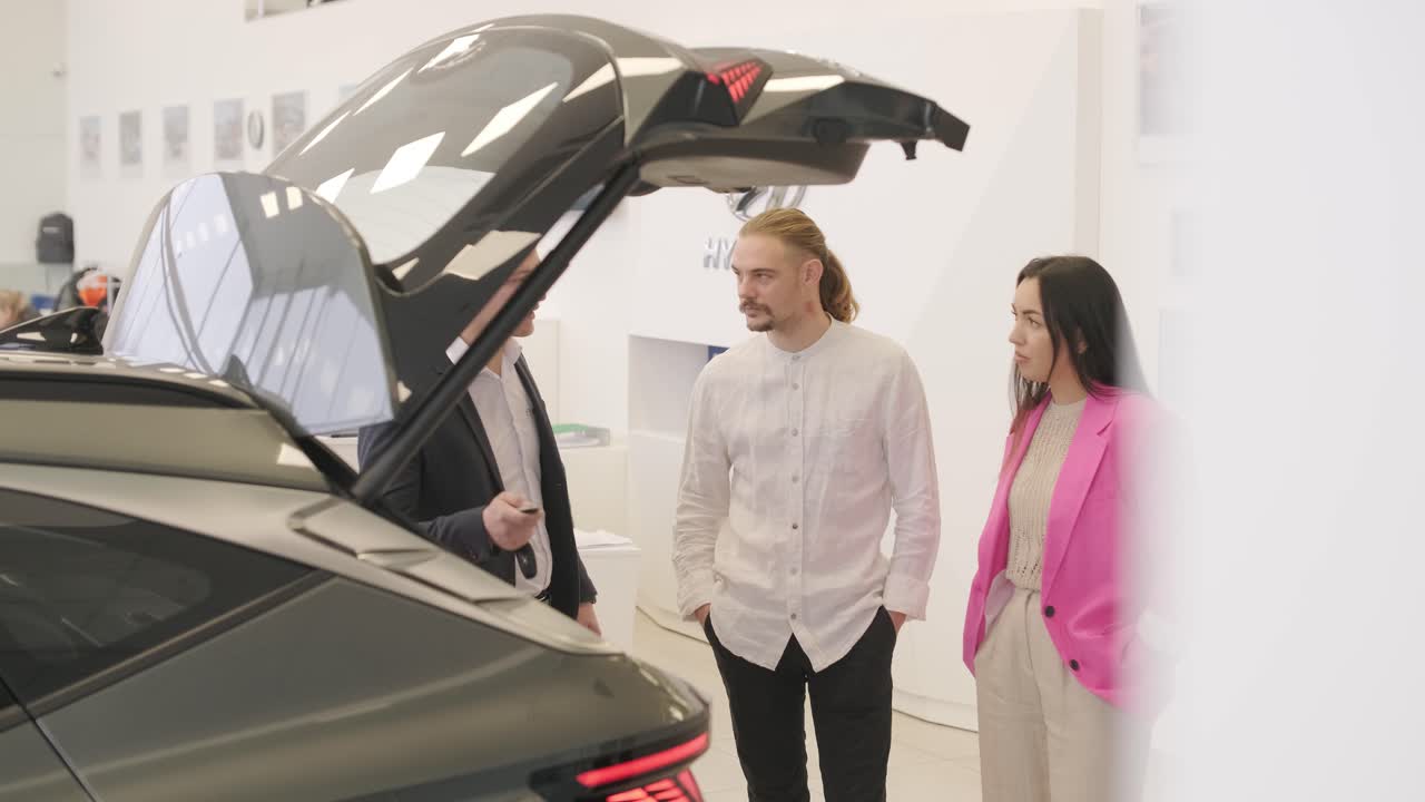 un joven vendedor de coches mostrando a una pareja de jóvenes un nuevo automóvil en el salón de concesionarios.