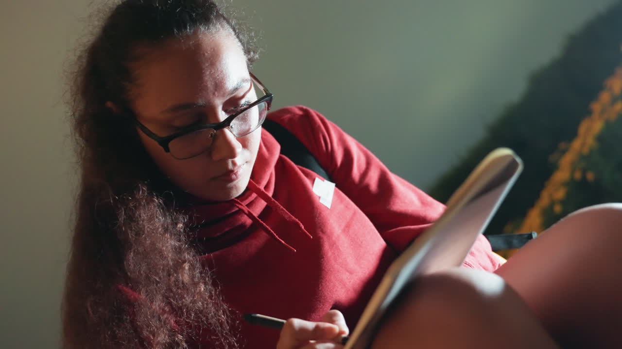 Person writing in notebook with glasses, red sweater and long curly hair, concentrated expression, indoor warm light, journal on lap, thoughtful composition, studying or drafting, candid portrait