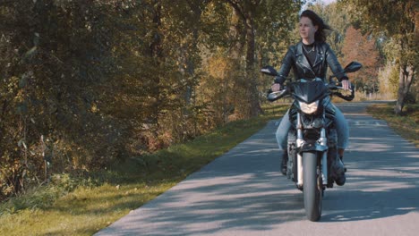 Hübsche-Lächelnde-Europäische-Junge-Frau,-Die-An-Einem-Sonnigen-Tag-Ein-Motorrad-Trägt,-Das-Lederjacke-Im-Wald-Mit-Lebhaften,-Bunten-Goldenen-Herbstblättern-Trägt