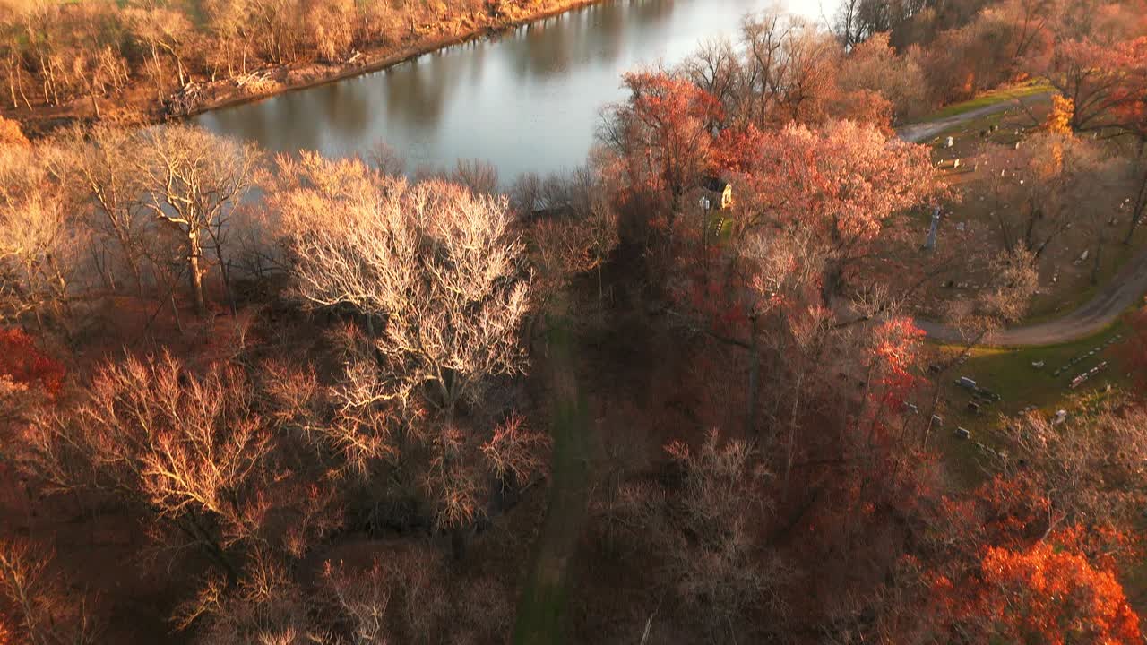 Drone view panning up from grass trail beside small creek to view of the river, autumn trees highlighted by sunset