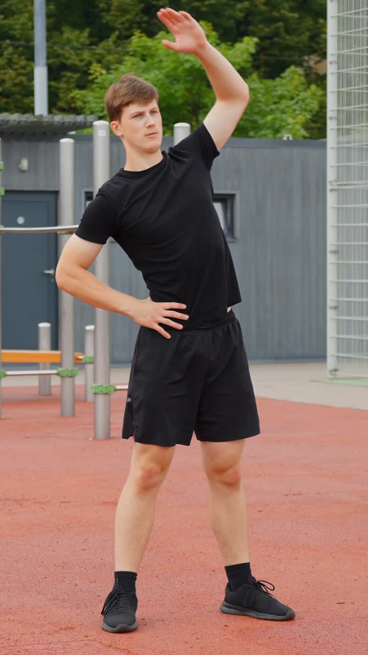 Young european man doing a side bend stretch with one arm raised and the other on his hip, wearing black athletic clothes while warming up for exercise outdoors, vertical static slow motion shot