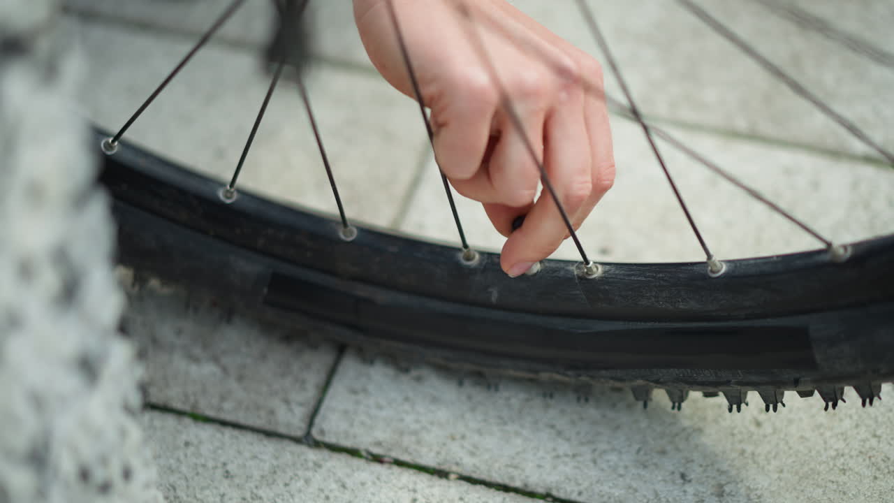 close de um pneu de bicicleta com uma mão removendo um espinho da lateral; mão feminina delicada puxa o espinho cravado da área do raio da roda no pavimento da cidade; banda de rodagem texturizada e válvula visíveis; clima de perseverança discreta