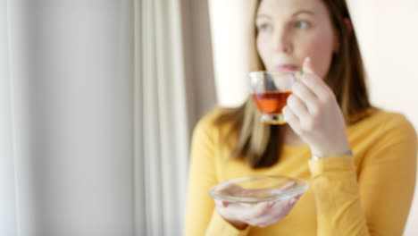 thoughtful caucasian woman drinking tea and looking through window at home, in slow motion