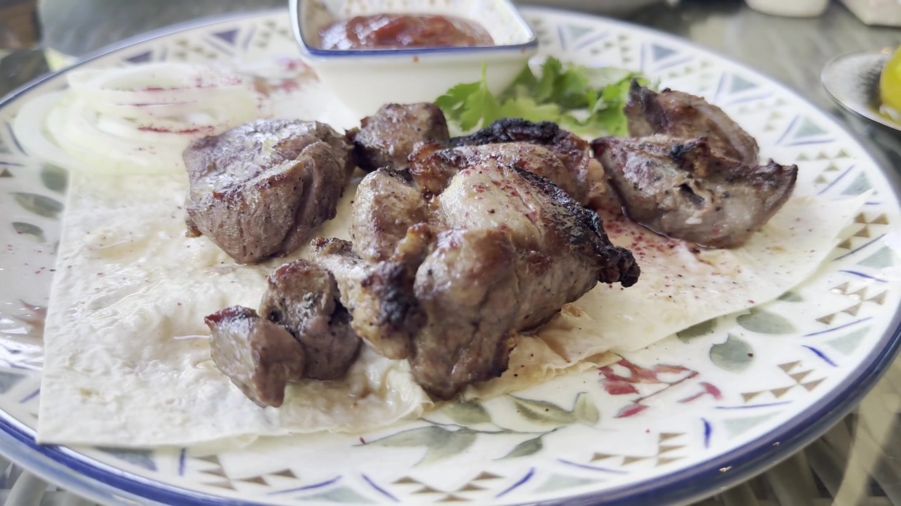 Un primo piano di un piatto di shashlik, spiedini di carne alla griglia, servito con pane lavash, cipolla rossa, sommacco, salsa di pomodoro ed erbe fresche.