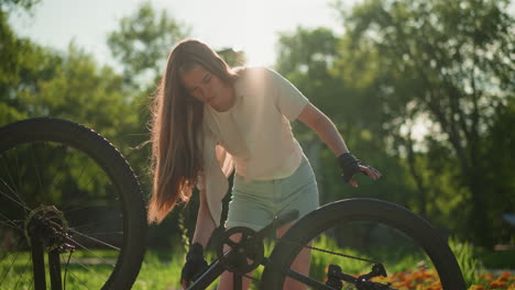 una joven se inclina ligeramente para girar el pedal de la bicicleta al revés, el cabello largo fluye hacia abajo, la luz del sol se refleja en ella mientras ajusta el neumático trasero, con árboles verdes exuberantes suavemente borrosos en el fondo